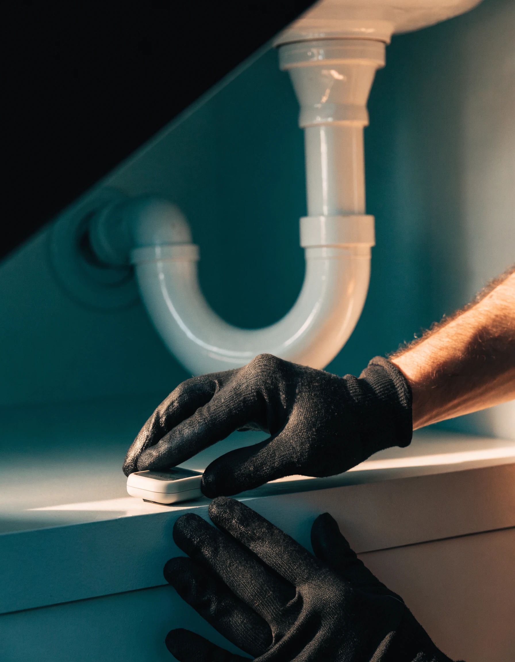A plumber in dark workwear inspecting pipework beneath a kitchen sink